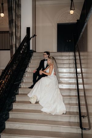 A couple sits together on grand marble stairs in an elegant venue with vintage lighting and architecture.