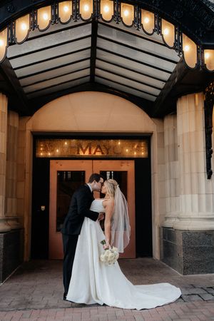 A wedding couple stands in front of an illuminated entrance with glowing pendant lights overhead.