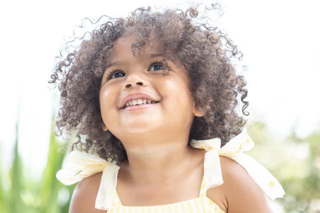 A smiling child with curly hair plays outdoors on a sunny day.