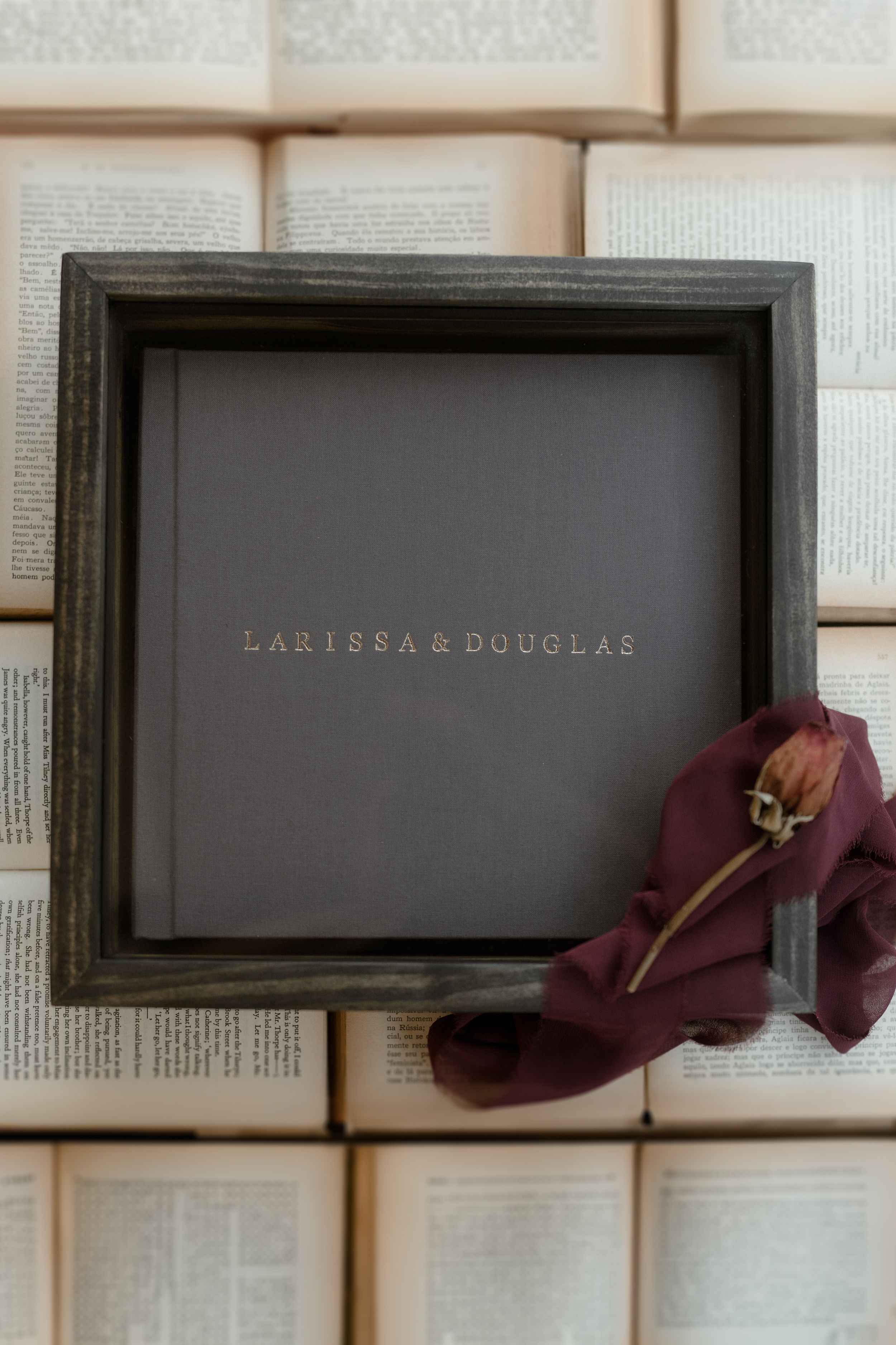 Elegant black photo frame with gold text reading 'LARISSA & DOUGLAS' displayed against book pages with dried flowers.