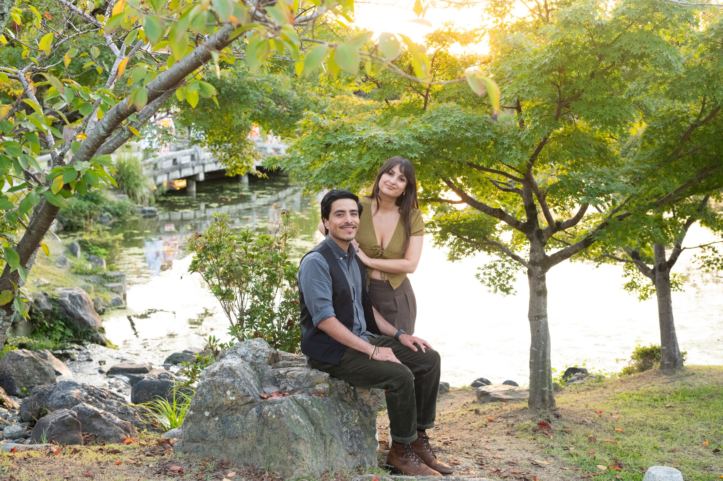 Couple sitting on rocks by a tranquil pond surrounded by lush green trees in warm afternoon light.