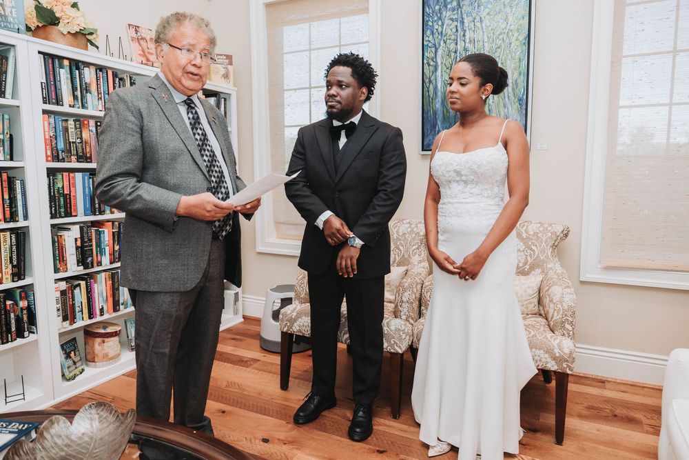 A couple stands with an officiant in a library setting during an intimate wedding ceremony.