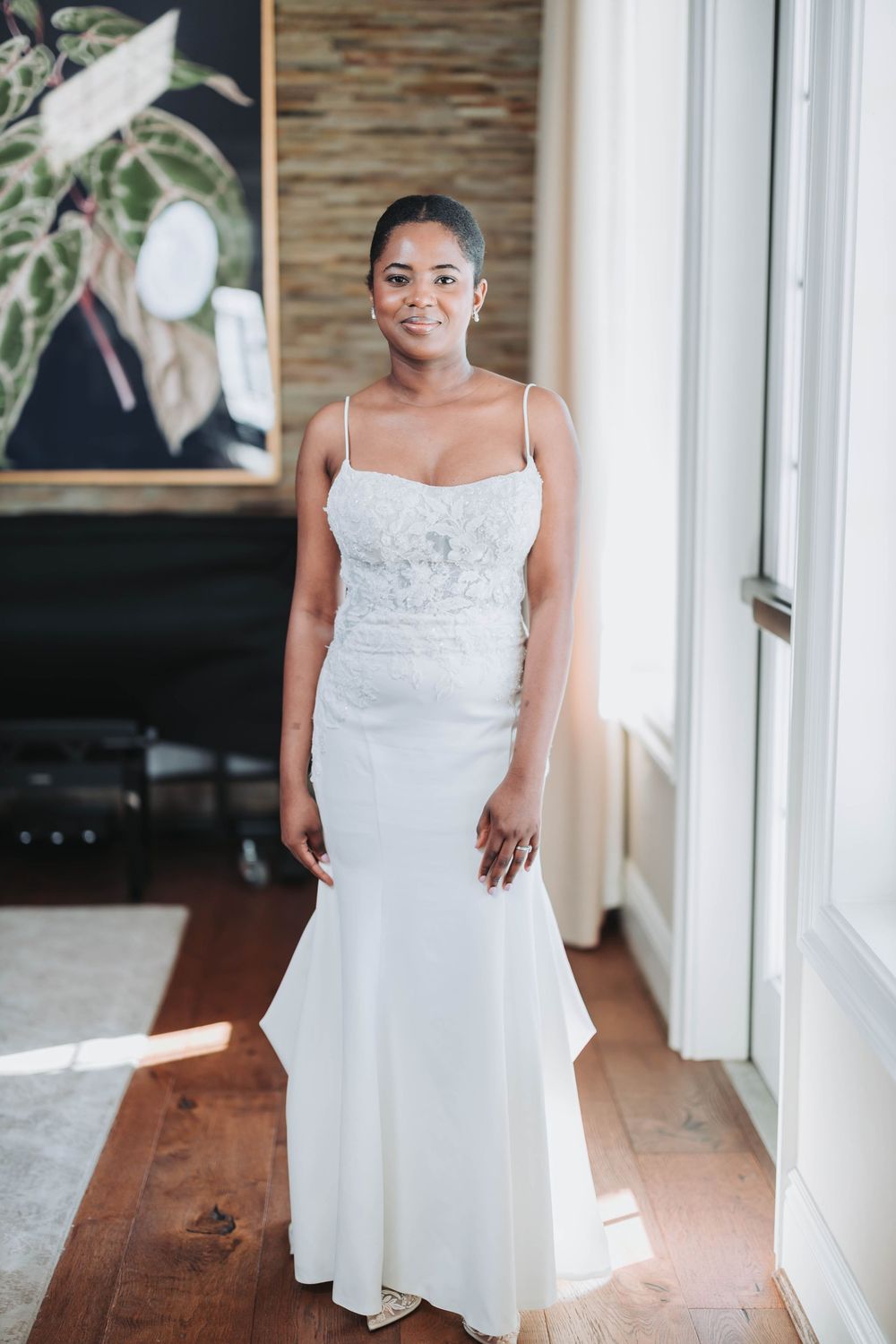 A bride in a white lace wedding dress stands in a sunlit hallway with hardwood floors and white walls.