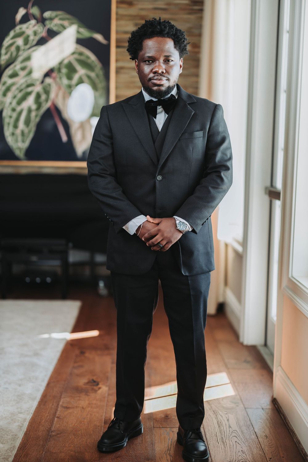 A groom in a black suit poses in a hallway with wooden floors and large windows.