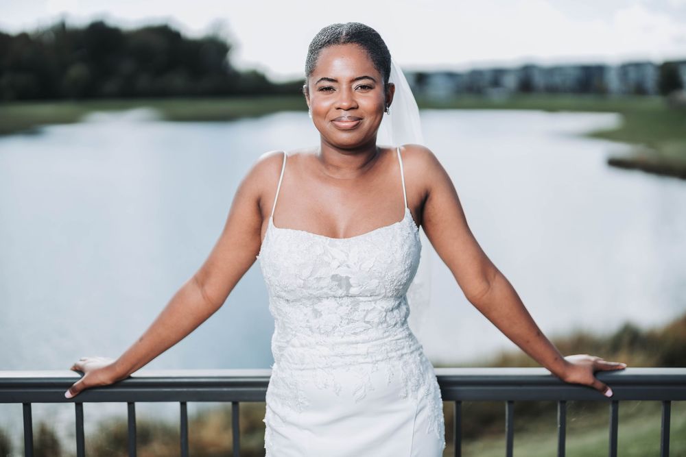 A person in a white wedding dress poses outdoors near a waterfront setting.