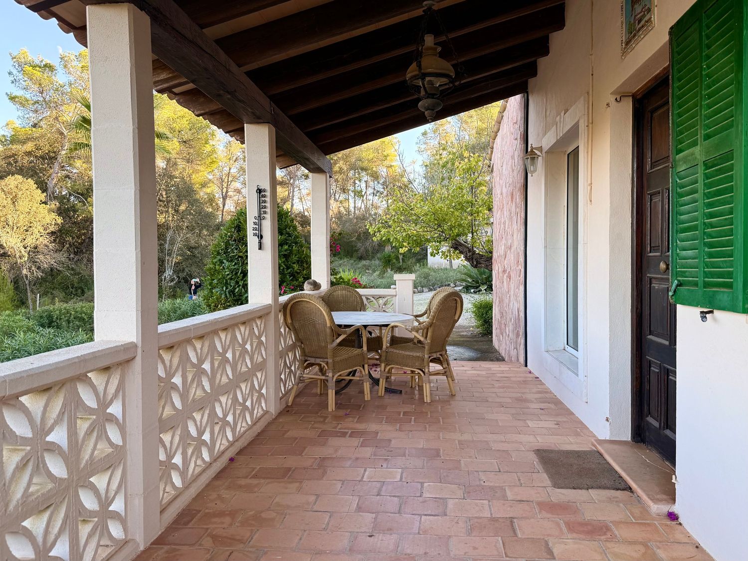 A long covered porch with terracotta tiles and green shutters features ornate white railings and outdoor seating.