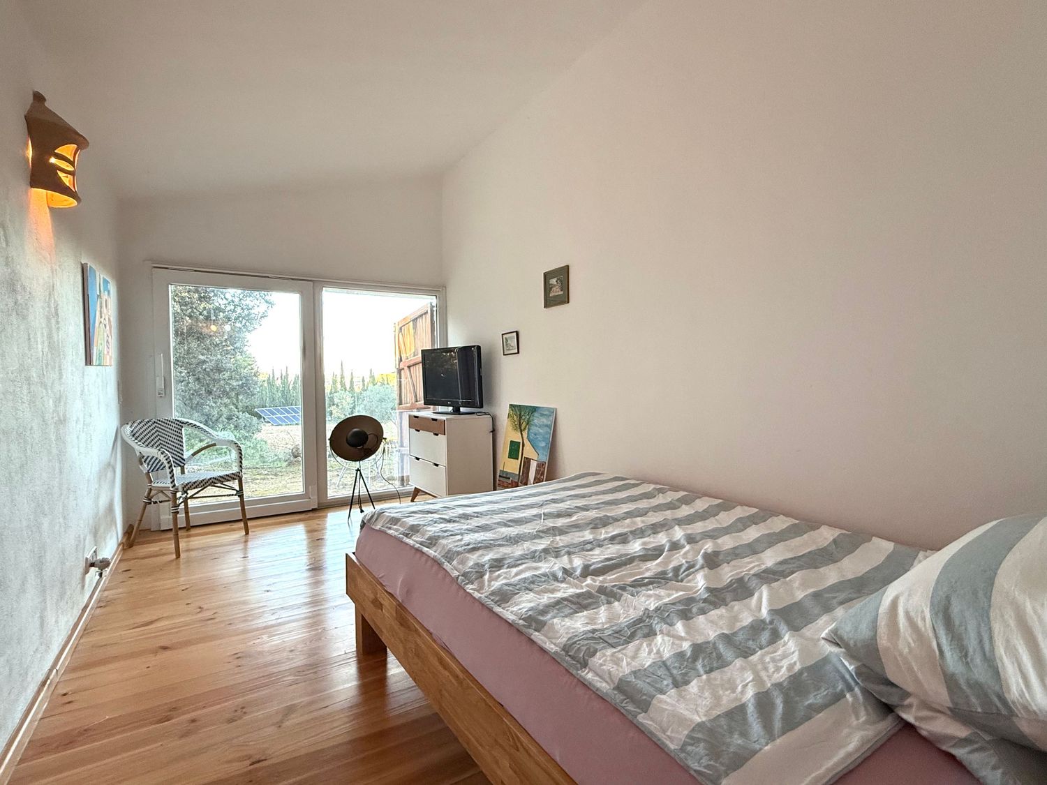 A modern bedroom interior with light wooden floors, white walls, and natural sunlight streaming through large windows.