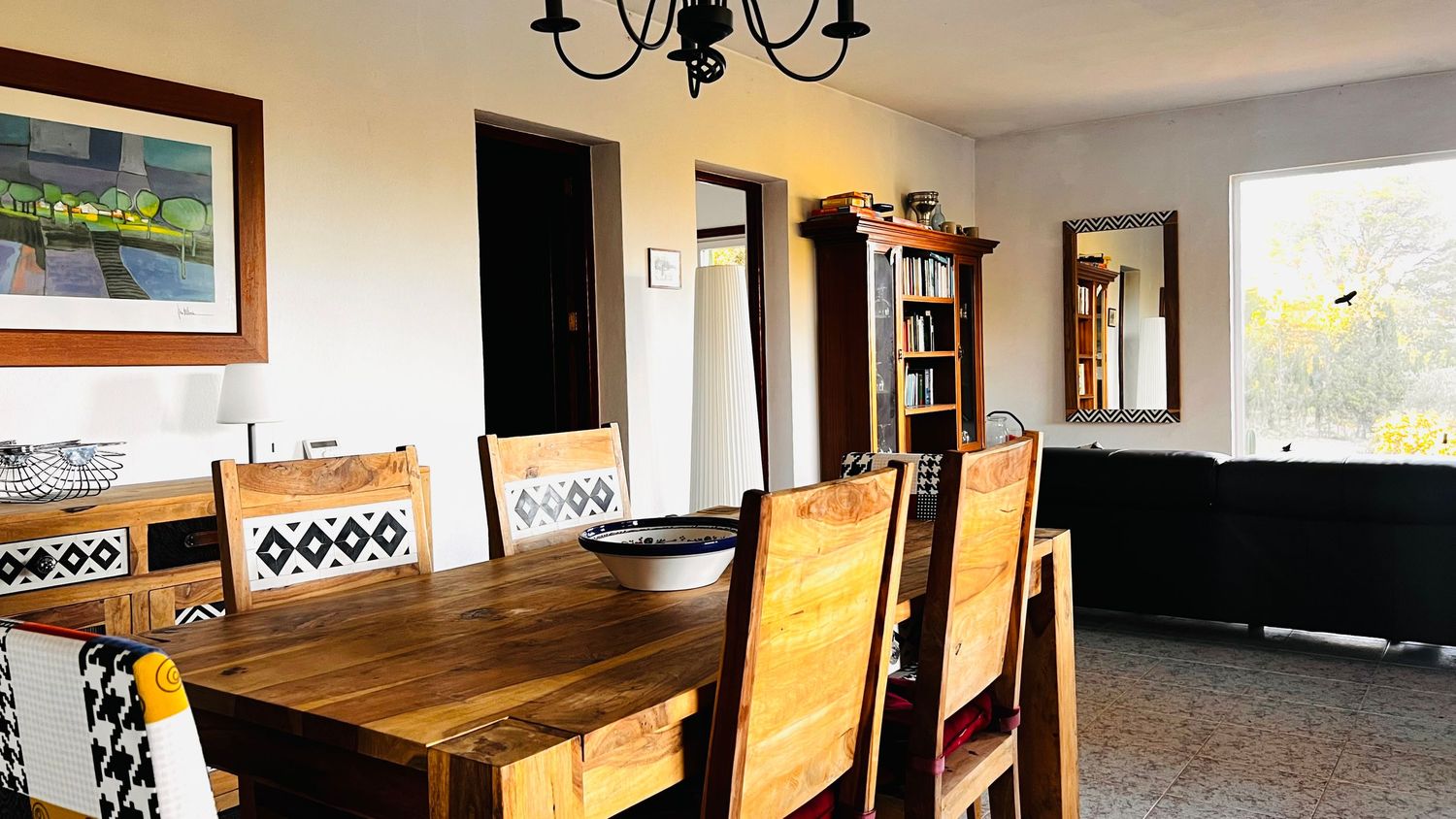A modern open-concept living space with wooden furniture, chandelier lighting, and large windows providing natural light.
