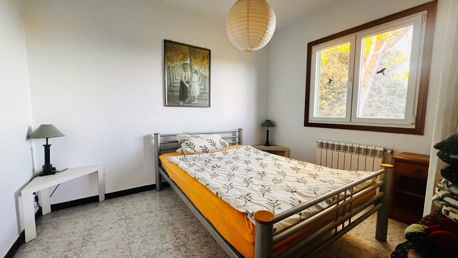 Simple bedroom with orange bed frame, floral bedding, white walls, paper lantern light fixture and framed artwork.