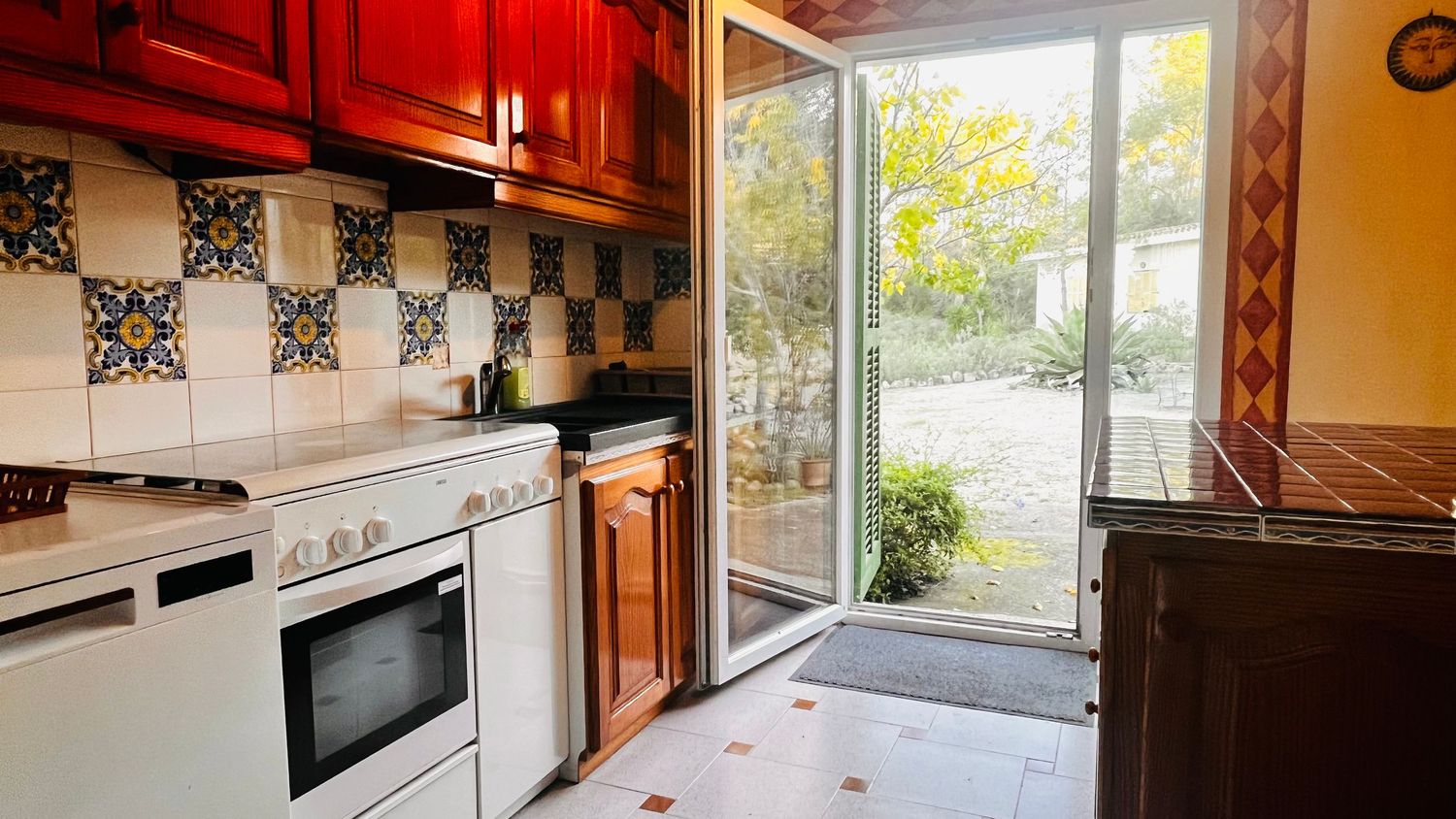 Modern kitchen with red cabinets, checkered backsplash, white appliances and glass door leading to garden view.