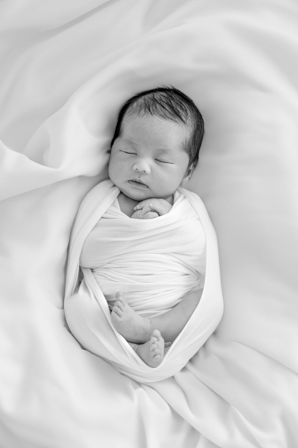 A sleeping newborn baby swaddled in white fabric against a soft white background.