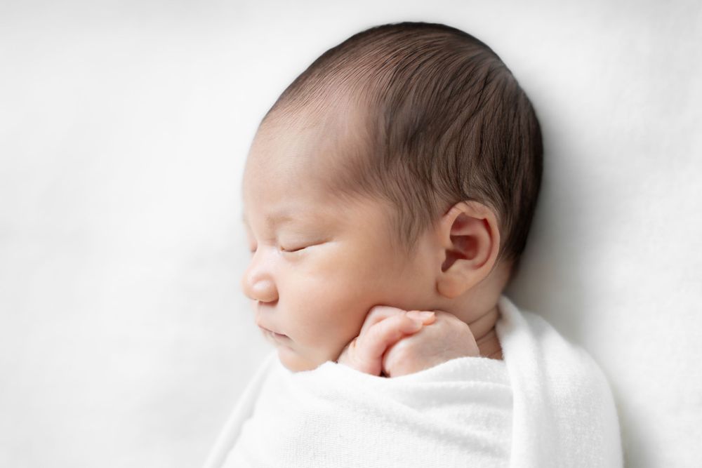 A peaceful sleeping newborn wrapped in soft white swaddling cloth against a light background.