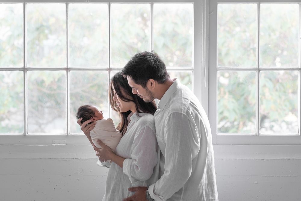 A couple in white clothing embraces tenderly by a bright window in a romantic moment.