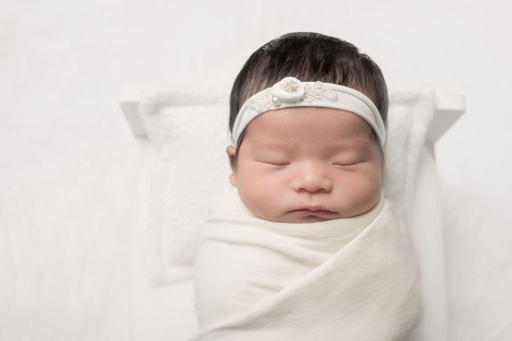 A sleeping newborn baby swaddled in white blankets with a white headband rests peacefully.