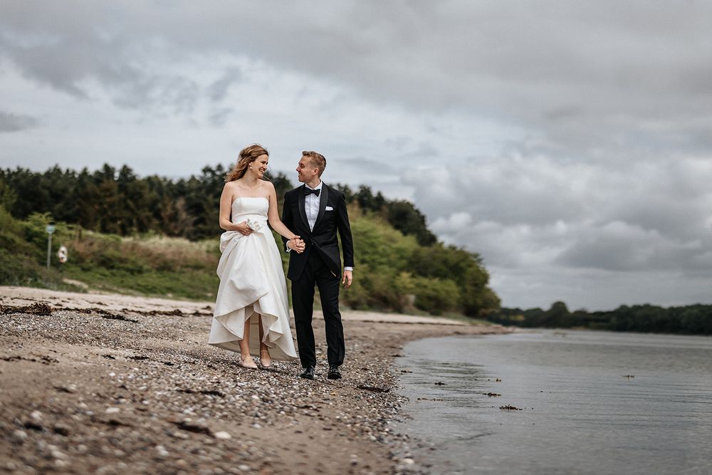 Couple in wedding attire walking together on a rocky beach shoreline with dramatic cloudy skies overhead.