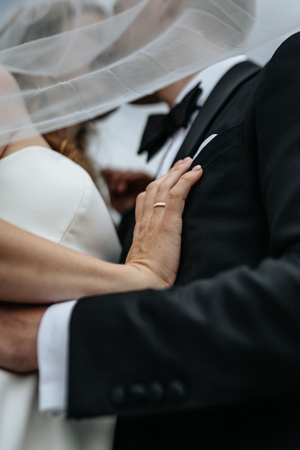 Close-up of newlywed couple embracing, showing white wedding dress, black tuxedo and delicate veil details.
