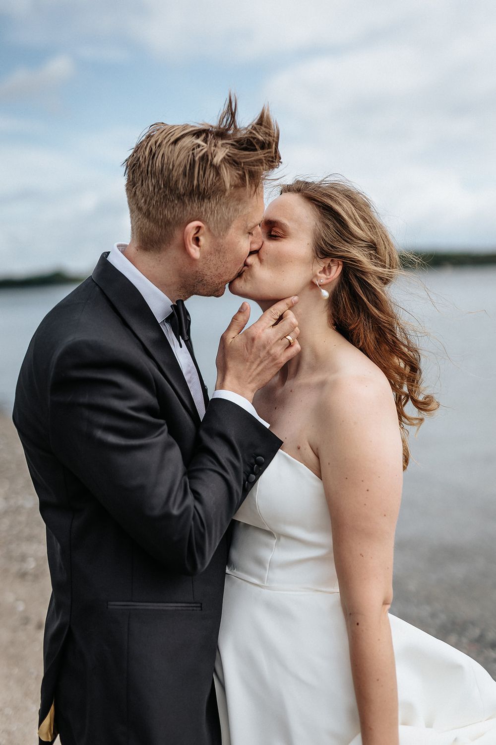 A romantic wedding couple shares a tender kiss by the water with windswept hair on their special day.