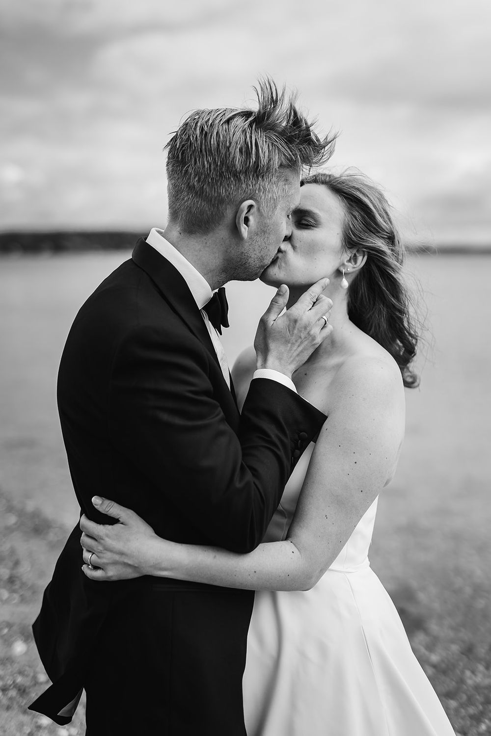 A romantic black and white wedding photo of a couple sharing a tender kiss by the water.