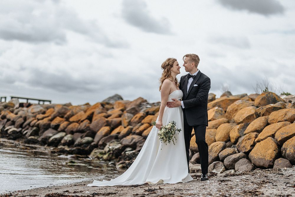 Wedding couple poses together on a rocky beach breakwater under dramatic cloudy skies.