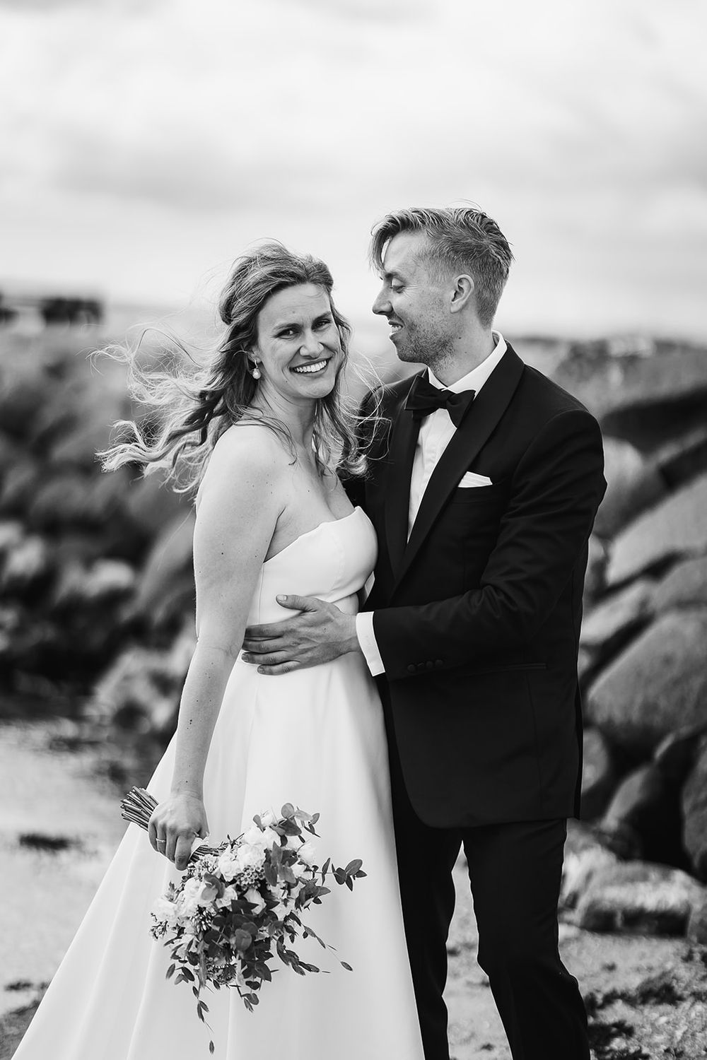 Black and white candid wedding portrait of newlyweds laughing together on a rocky shore.