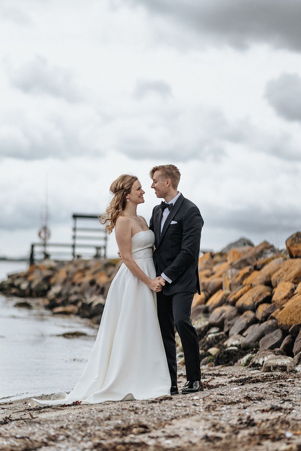 A couple in wedding attire stands together on a rocky beach shoreline under dramatic cloudy skies.