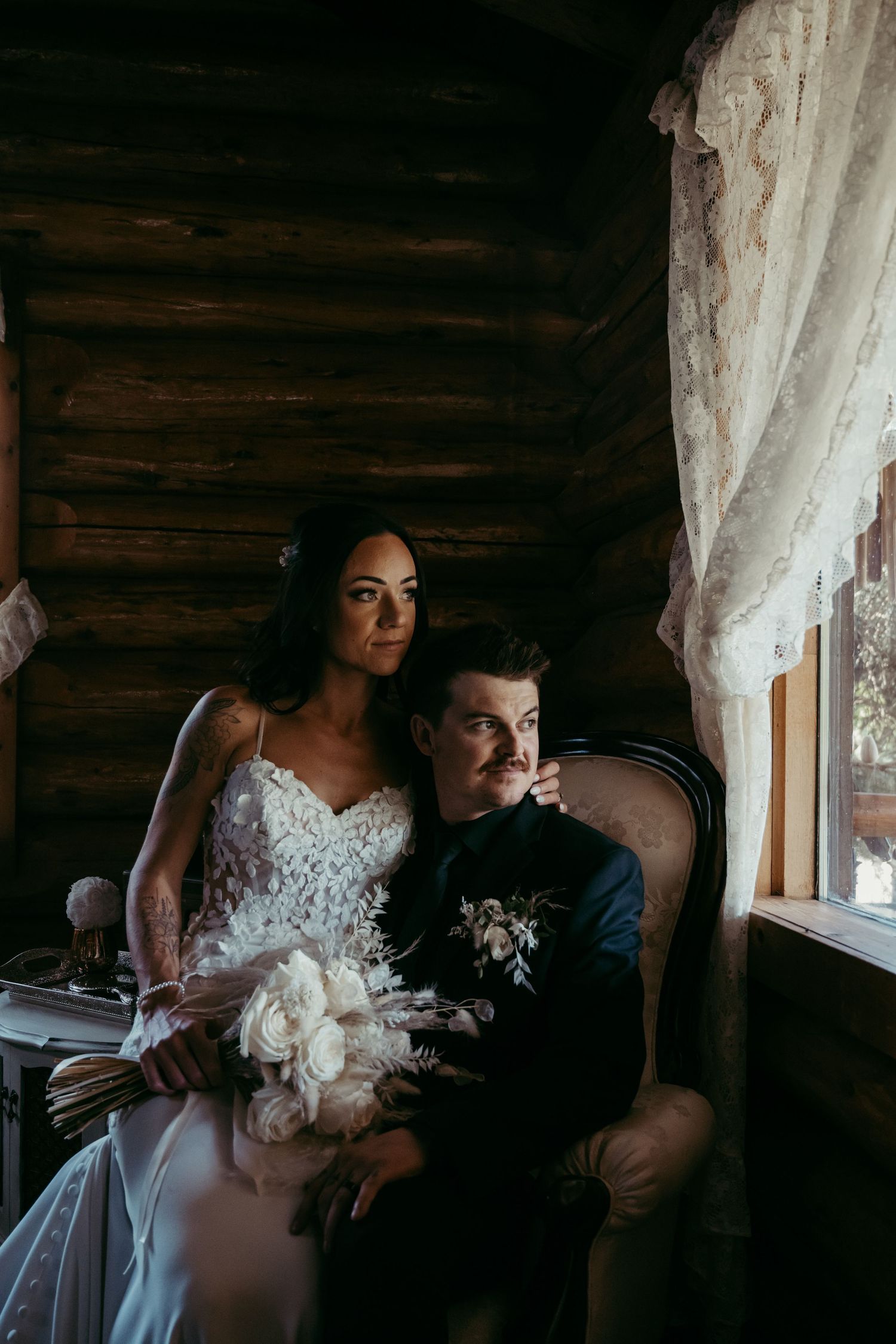 A bride and groom sit together in a rustic cabin interior with white lace curtains and natural window light.
