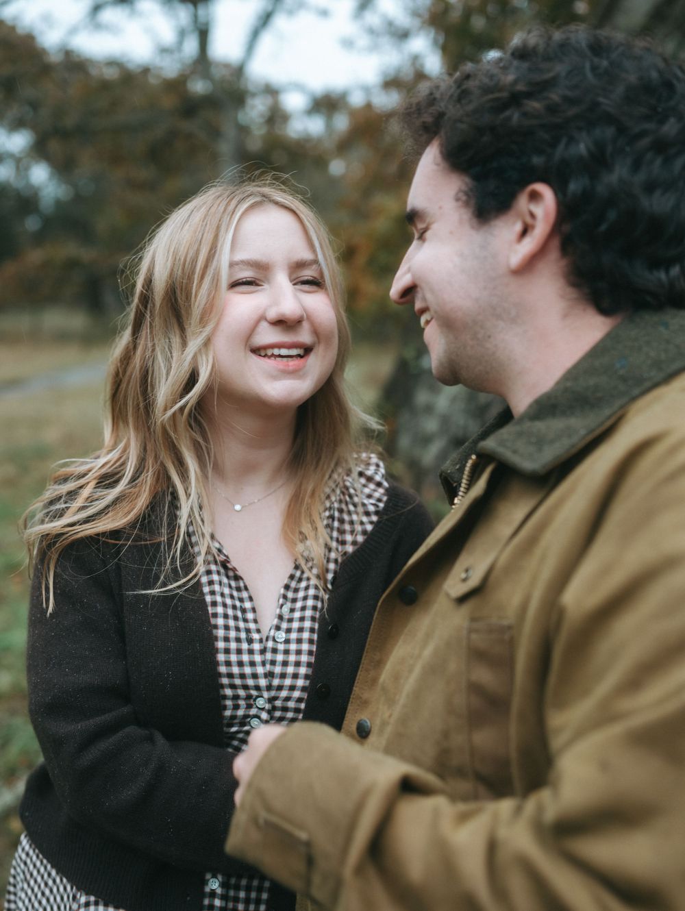 A couple laughs together in a natural outdoor setting with fall foliage in the background.