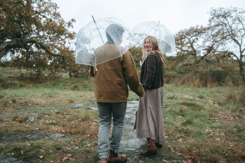 Two people stand together in a misty autumn field, one holding a clear umbrella, wearing casual outdoor attire.