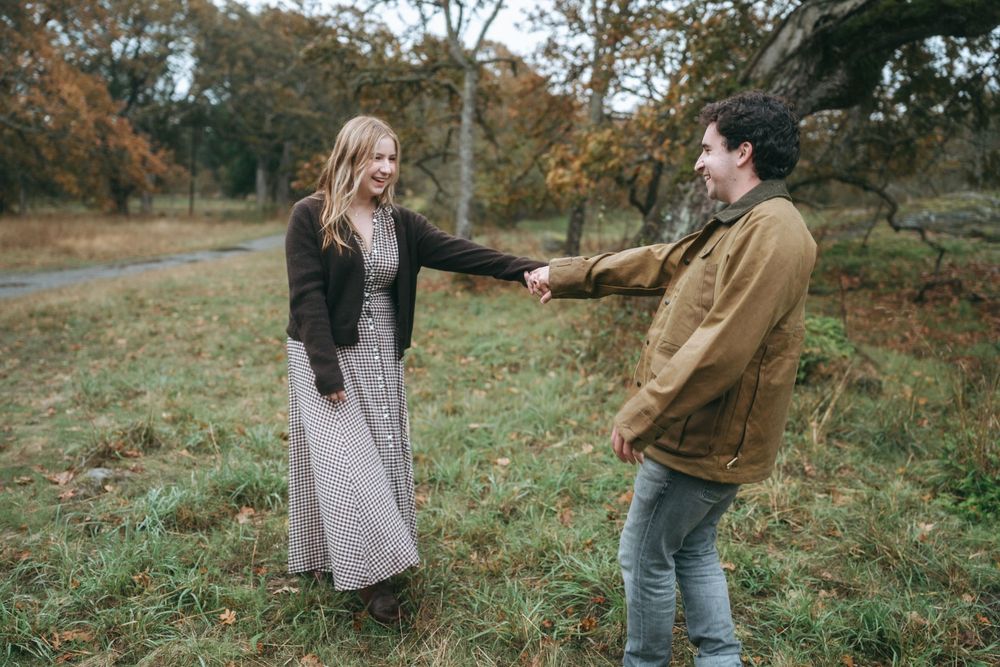 A couple playfully interacts in a scenic autumn field with brown and green foliage in the background.