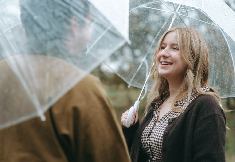 Person with blonde hair smiles while holding clear umbrella in rainy weather wearing black and white outfit.