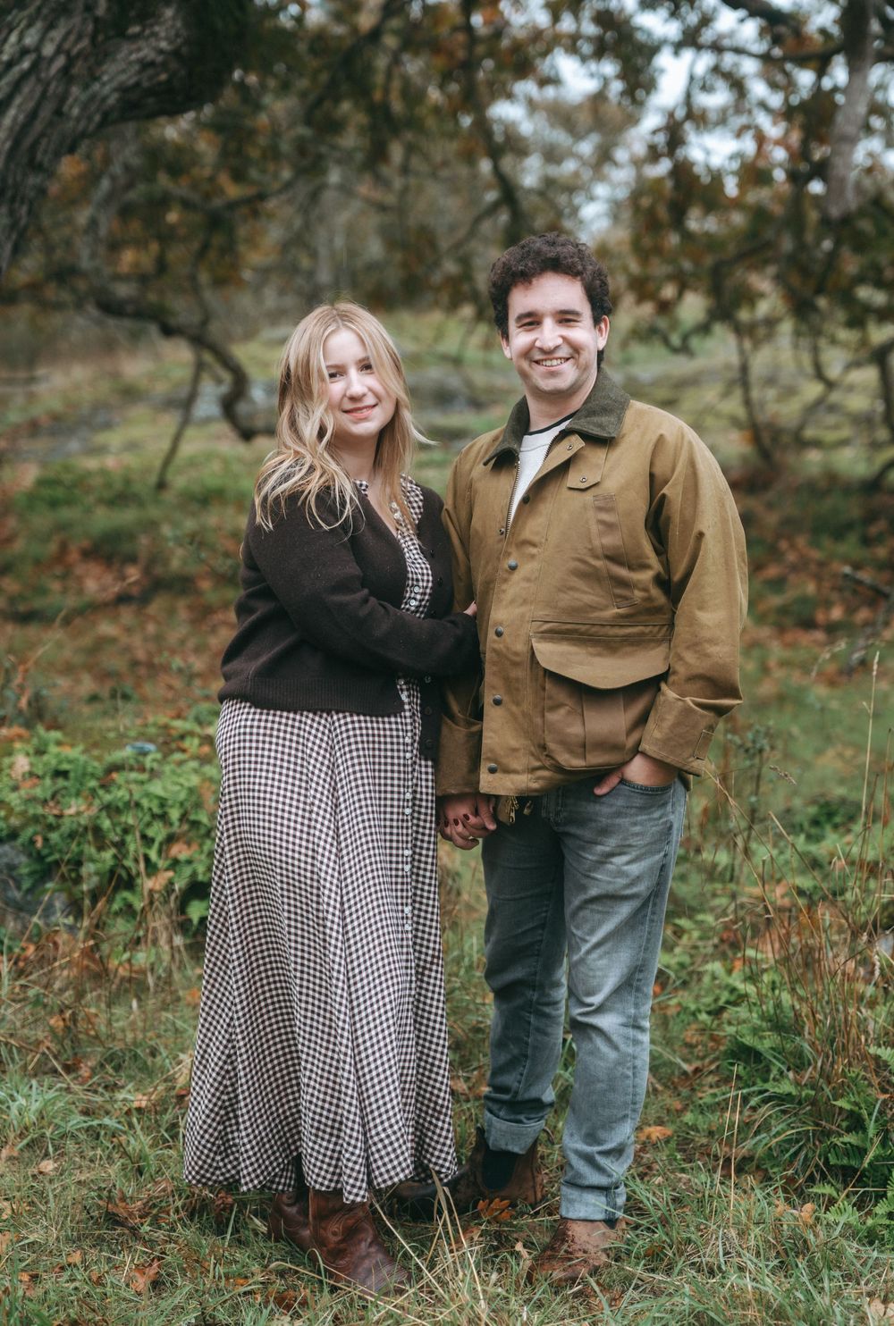 A couple poses together in an autumn forest setting wearing casual clothing and a checkered dress.