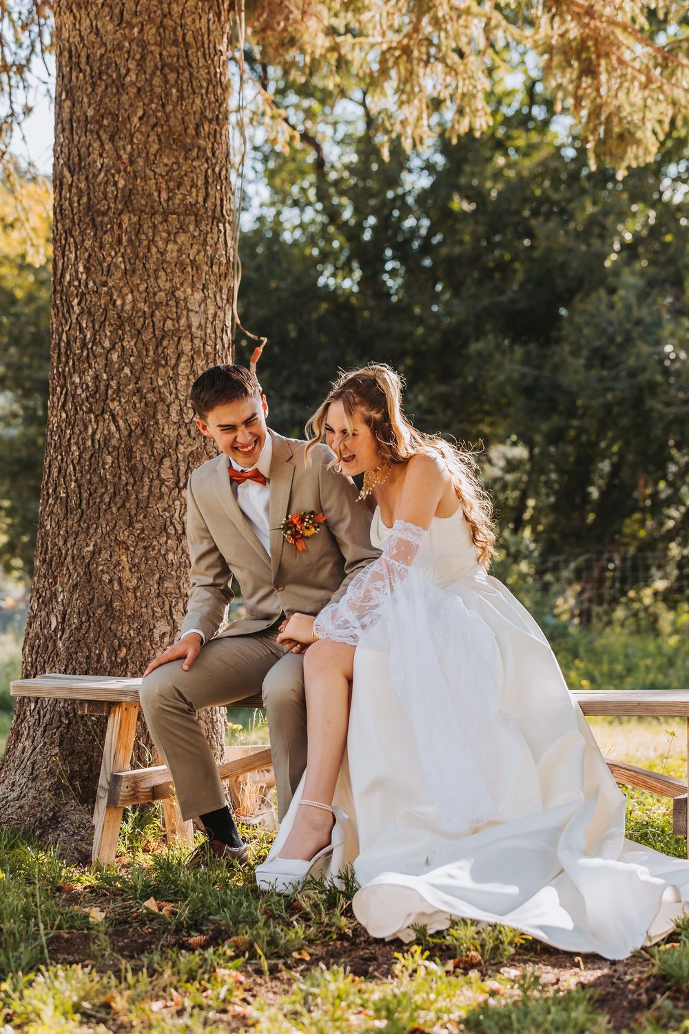A couple in wedding attire sits together under a tree on a sunny autumn day sharing candid laughter and joy.