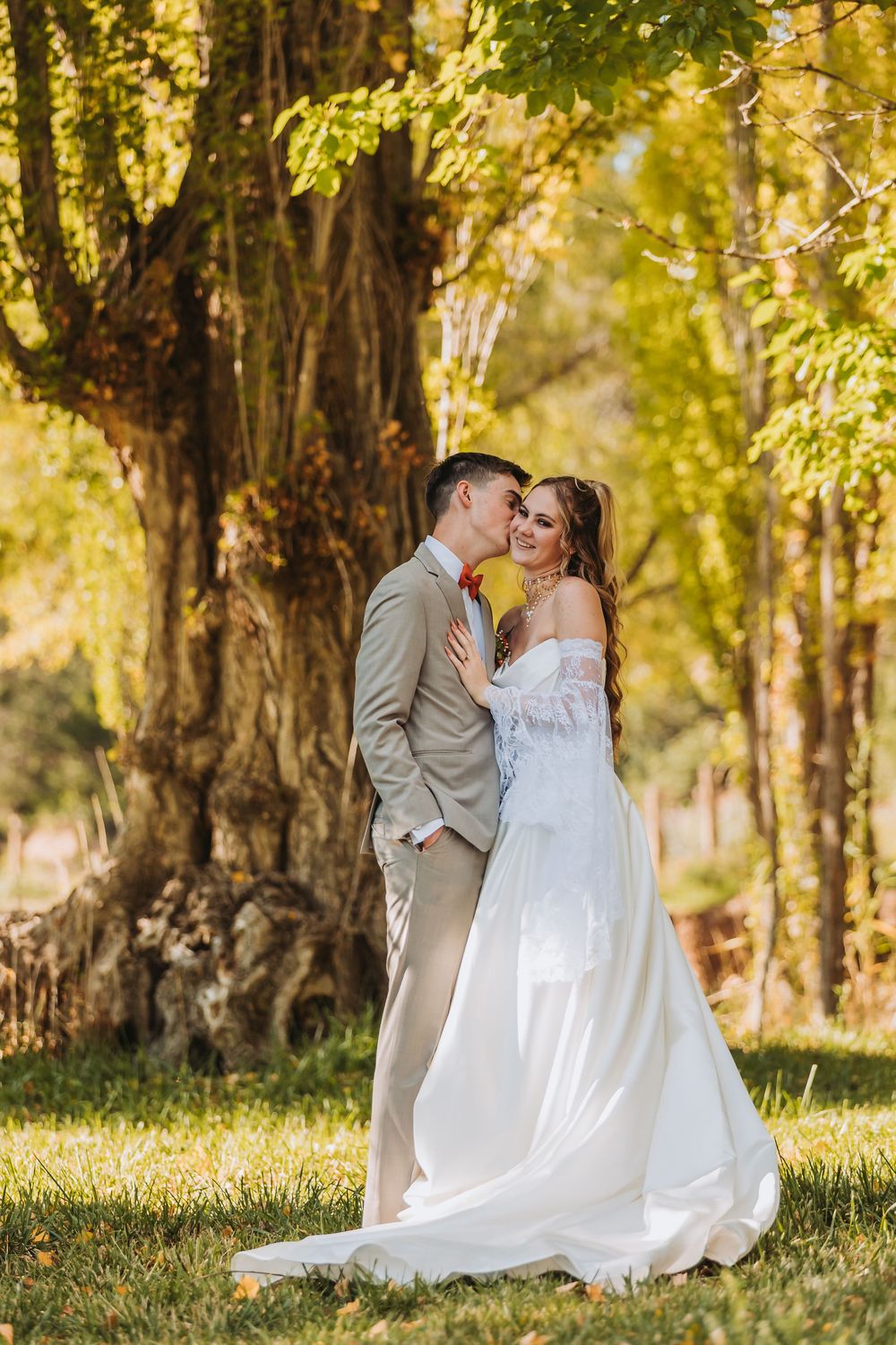 A romantic wedding couple poses together in a sunlit forest setting with tall trees and golden autumn light.
