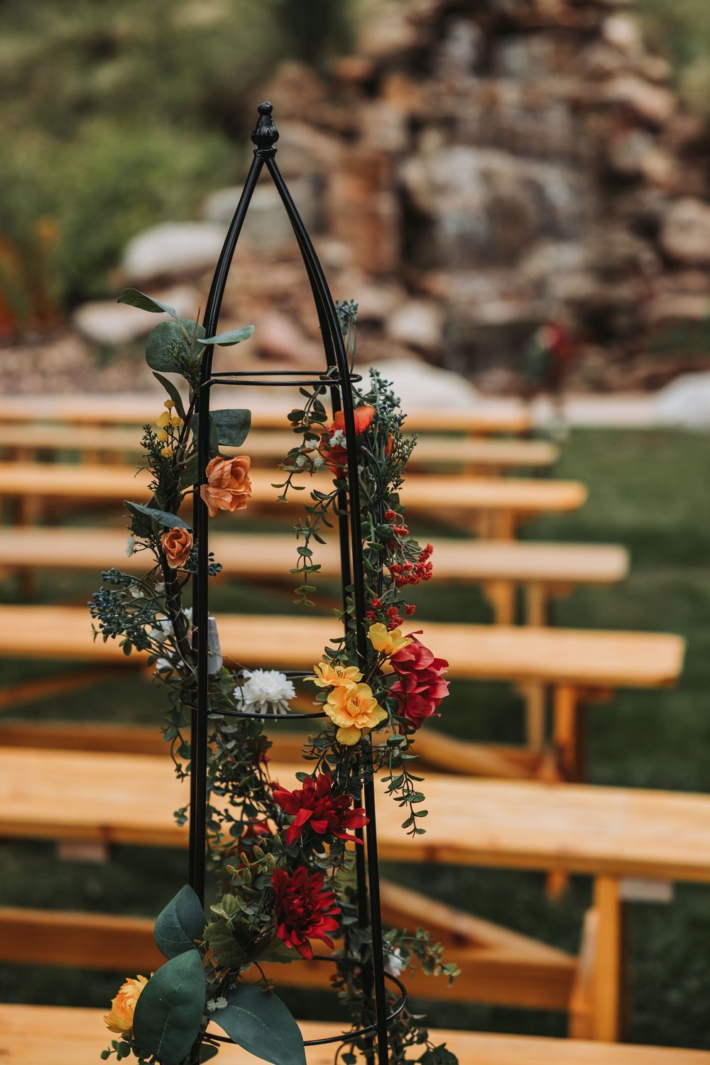 Black metal wedding arch decorated with colorful flowers and greenery stands beside wooden benches at outdoor ceremony.