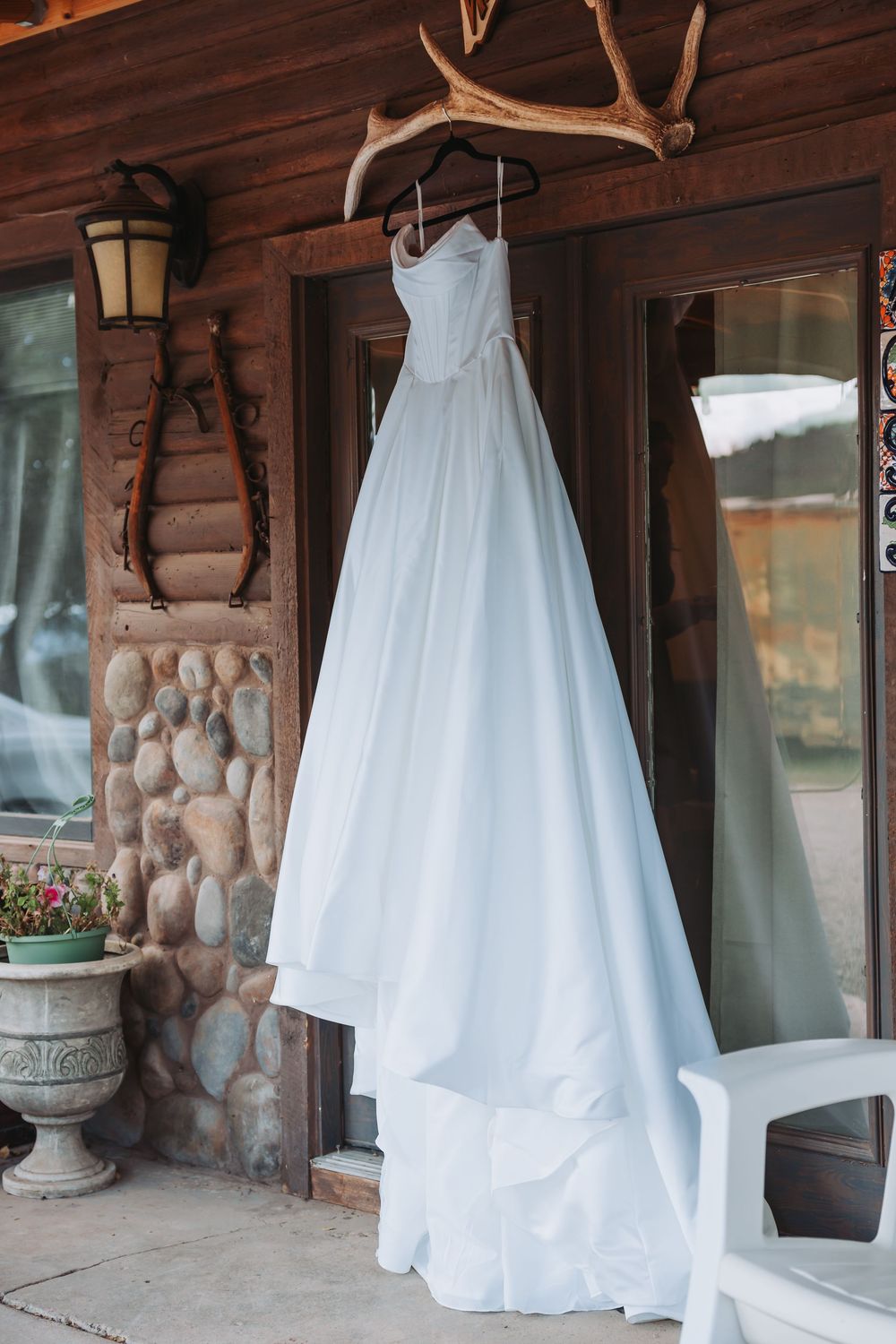 A white wedding dress hangs from antlers mounted on a rustic log cabin wall with stone accents and outdoor lighting.