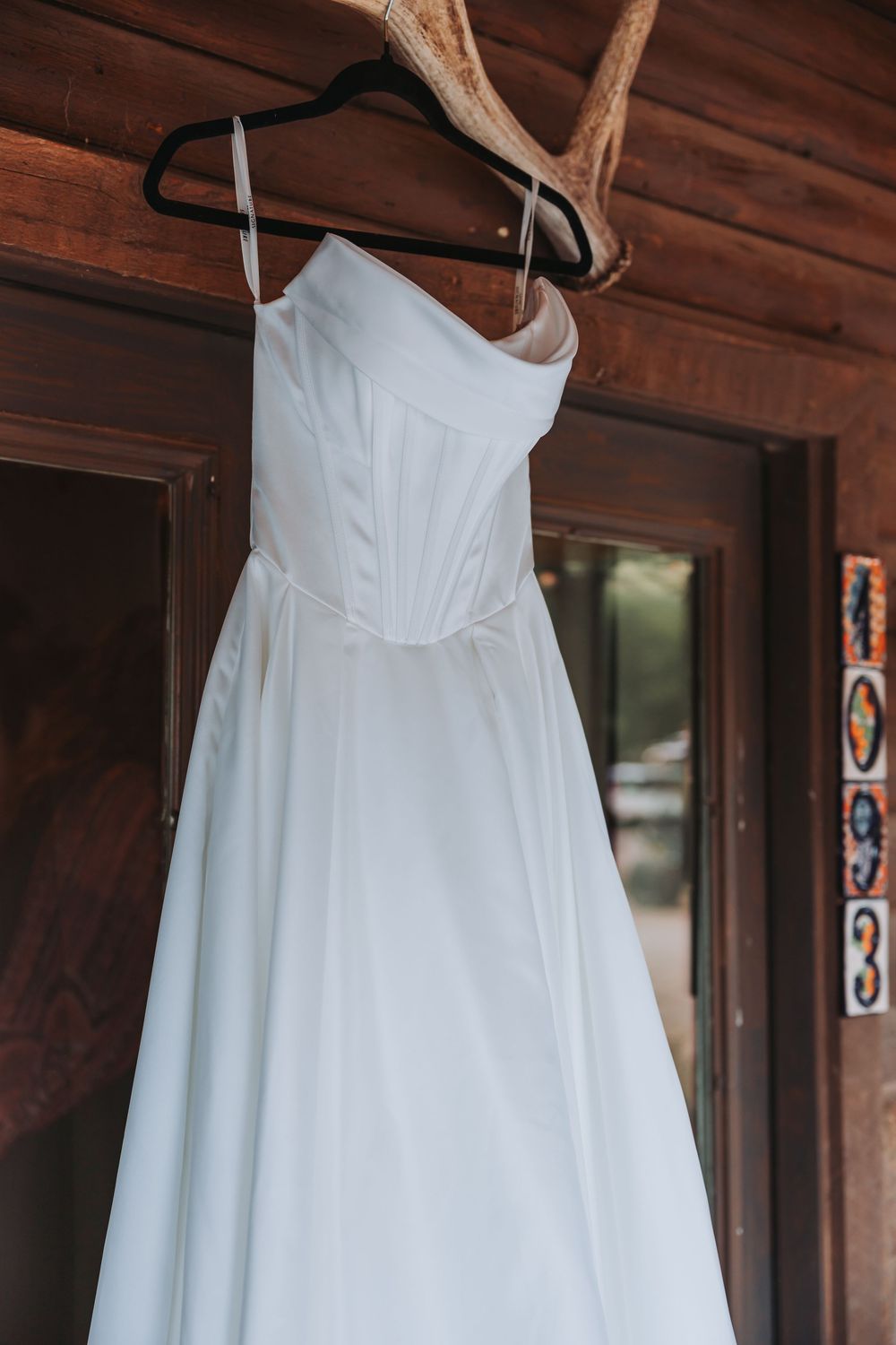 A white wedding dress with a fitted bodice and flowing skirt hangs on a black hanger against a rustic wooden wall.