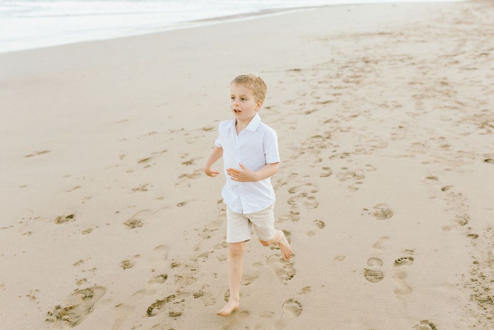 A child in white clothing runs barefoot along a sandy beach on a bright day.