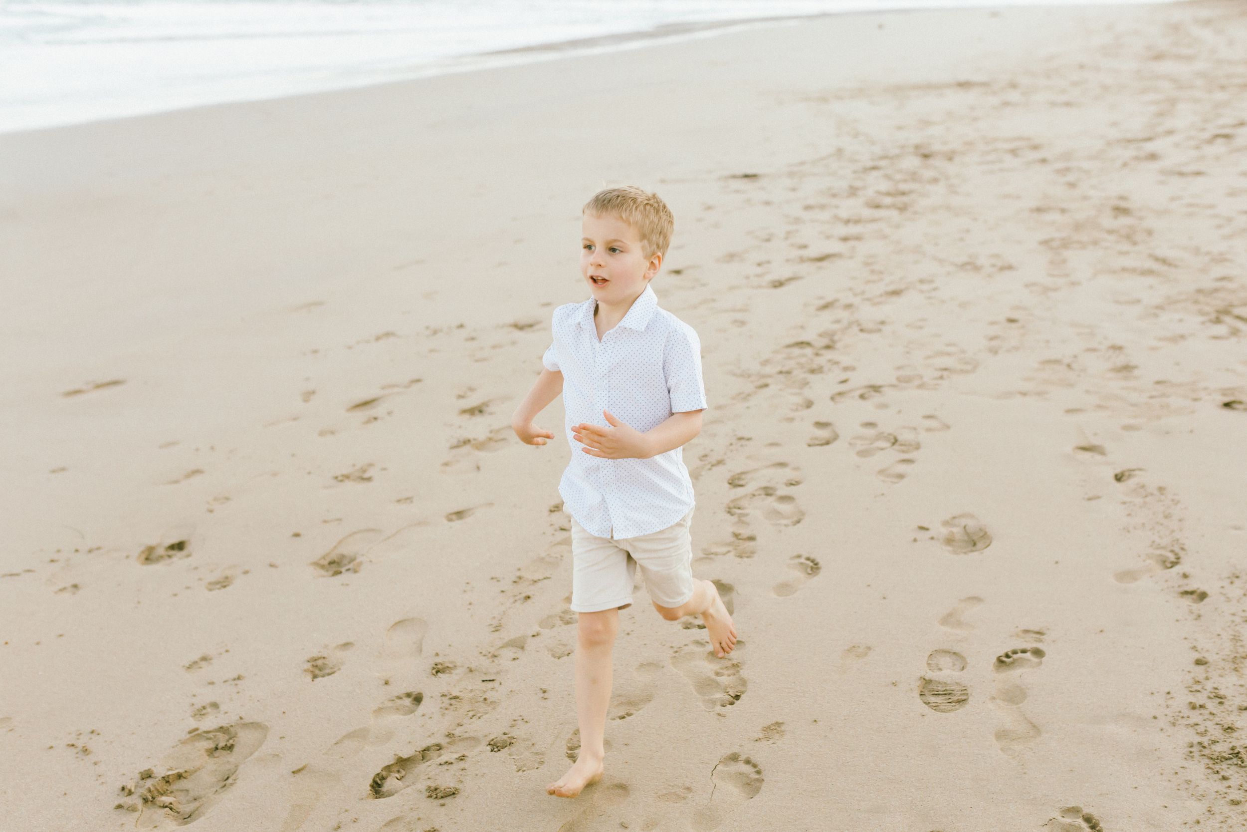 A child in white clothing runs barefoot along a sandy beach on a bright day.