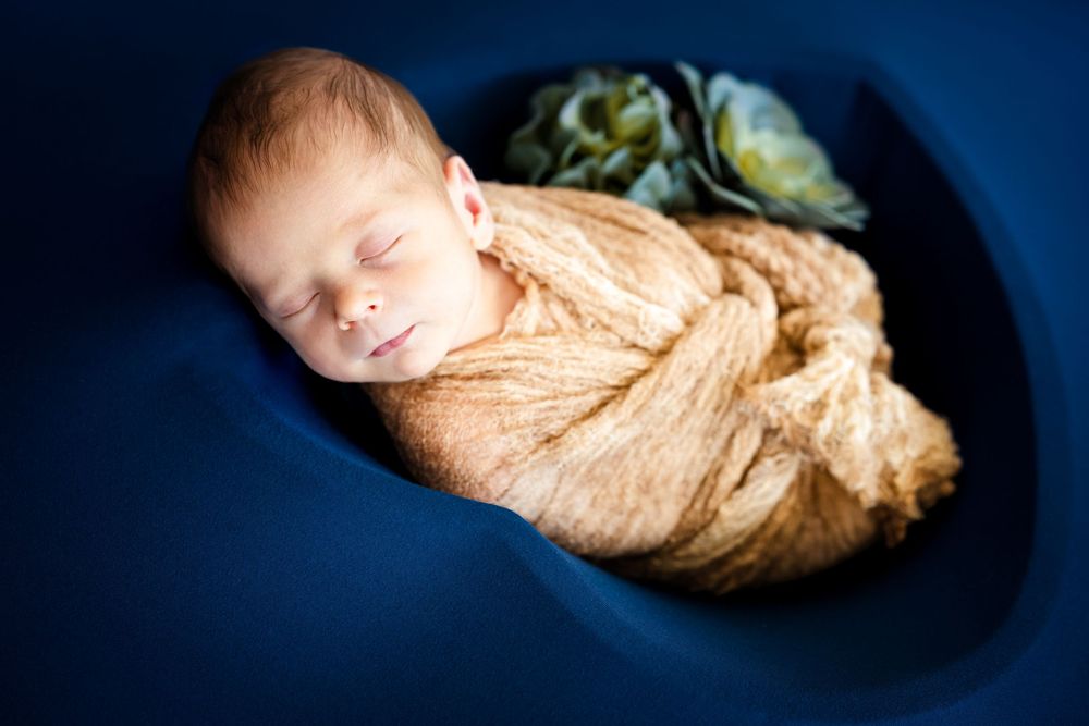 Peaceful newborn baby swaddled in a golden wrap, sleeping in a blue bowl with floral accents.