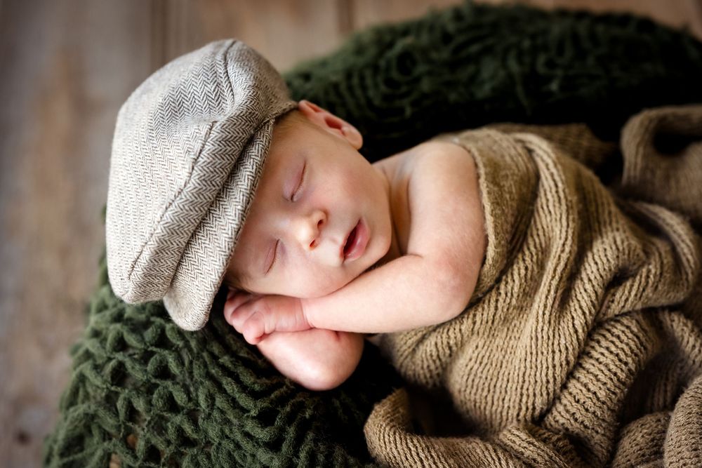 Sleeping newborn baby wearing a gray flat cap, wrapped in knit blankets in earthy tones.