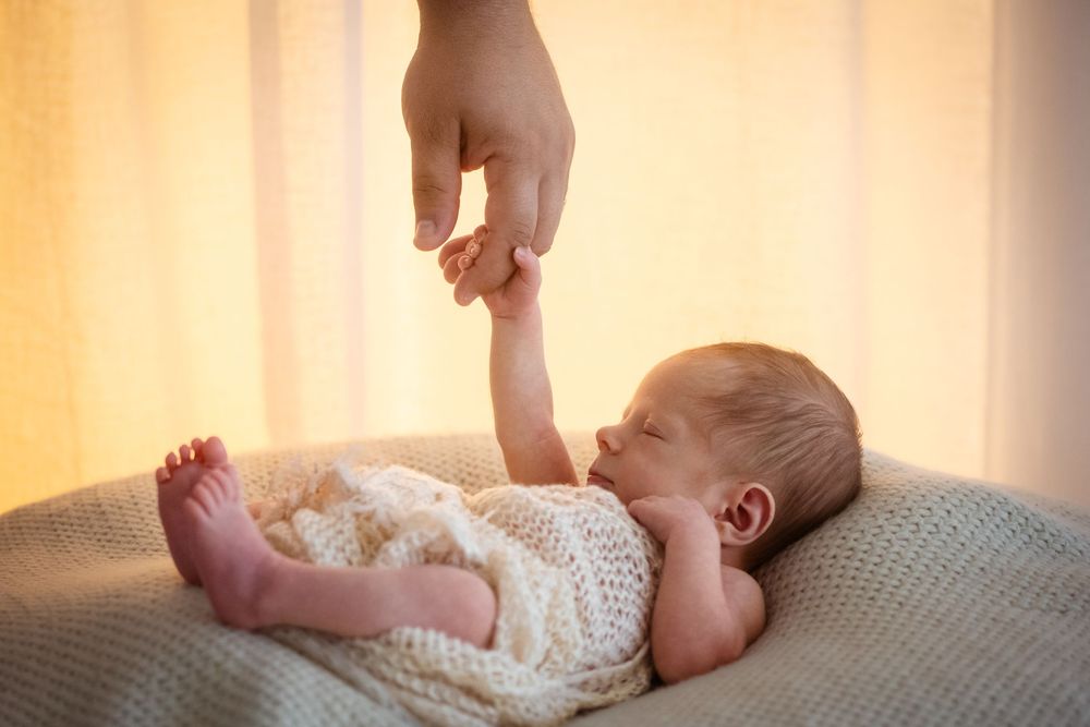 Newborn baby sleeping peacefully while gently holding an adult's finger, wrapped in a knit blanket.