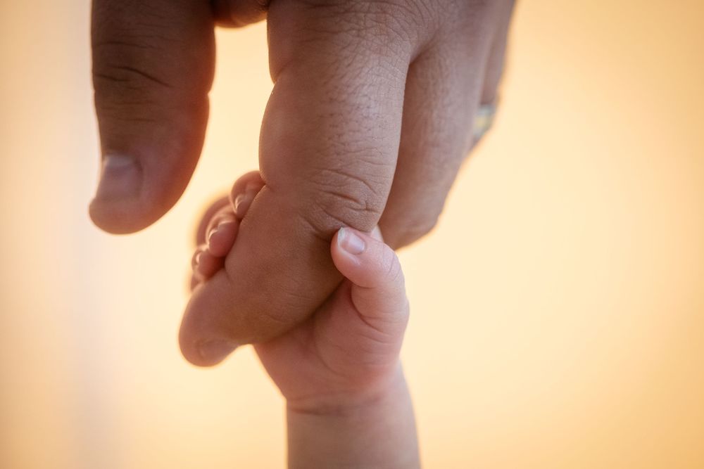 Close-up of a baby's tiny hand gripping an adult's finger against a warm beige background.