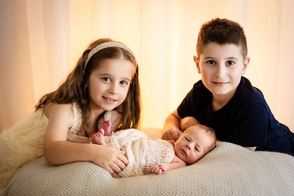 Two siblings smile proudly beside their sleeping newborn baby wrapped in pink knit, posed on a soft white blanket.
