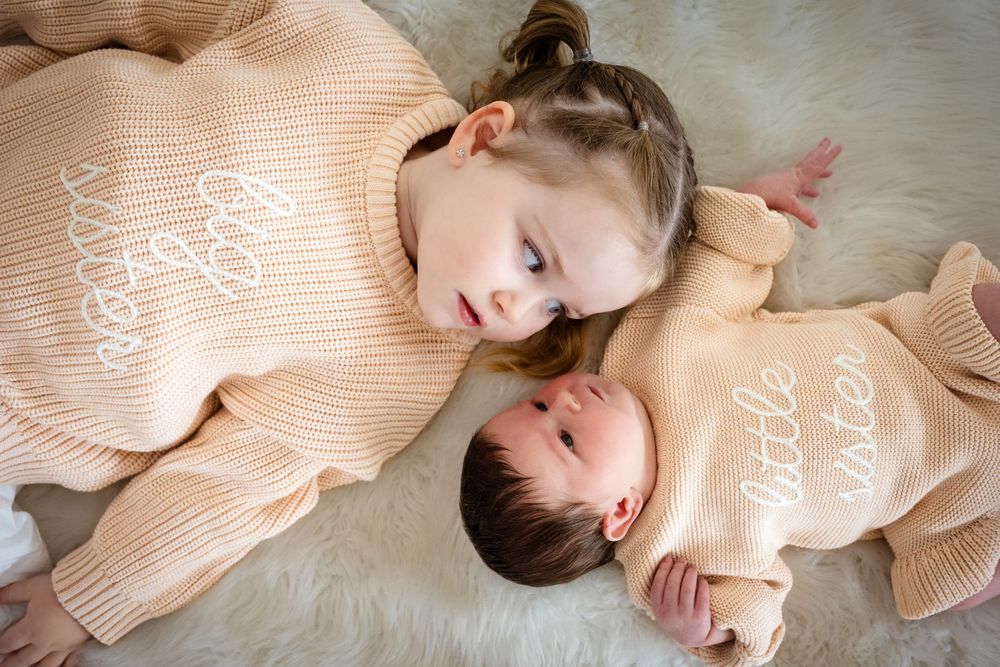 Two sisters in matching beige knit sweaters, older girl looking at newborn baby, lying on a fluffy white rug.