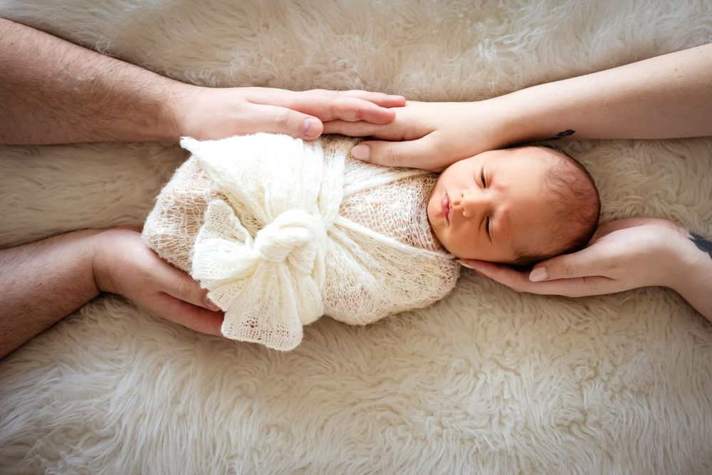 Newborn baby swaddled in white knit wrap, gently held by parents hands on soft cream fur blanket.