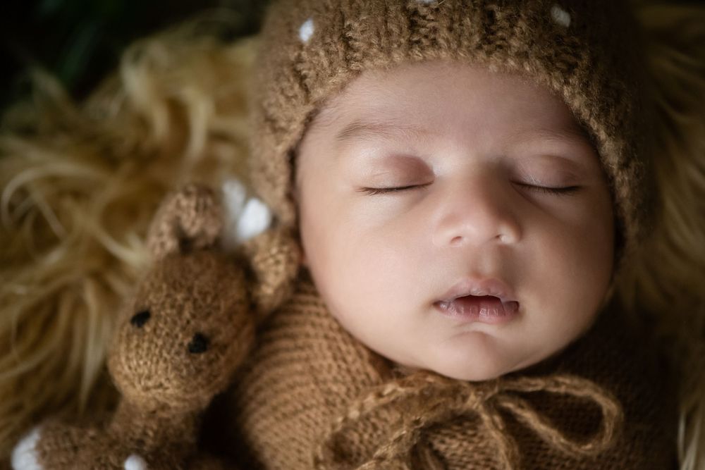 Sleeping newborn baby in brown knit hat and outfit cuddles with teddy bear on fluffy fur blanket.