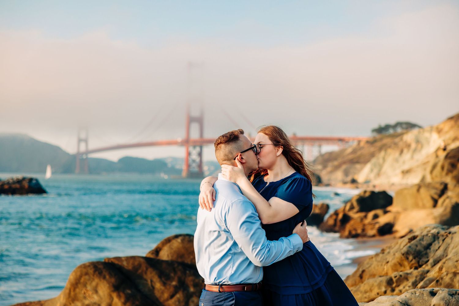 Romantic couple embraces on rocky coastline with Golden Gate Bridge visible in the misty San Francisco background.