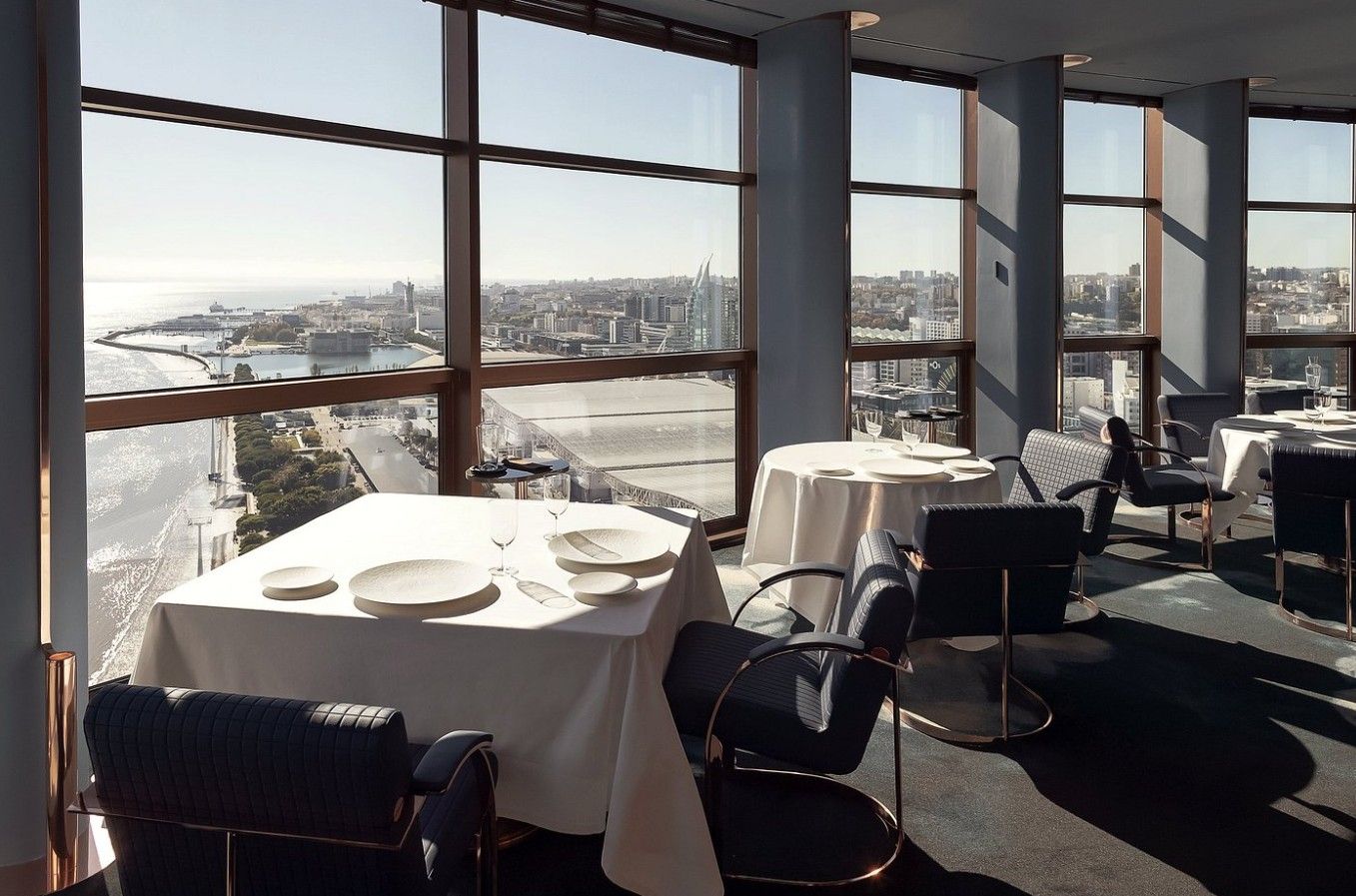 Elegant restaurant interior with panoramic windows overlooking the ocean, featuring white tablecloths and modern decor.