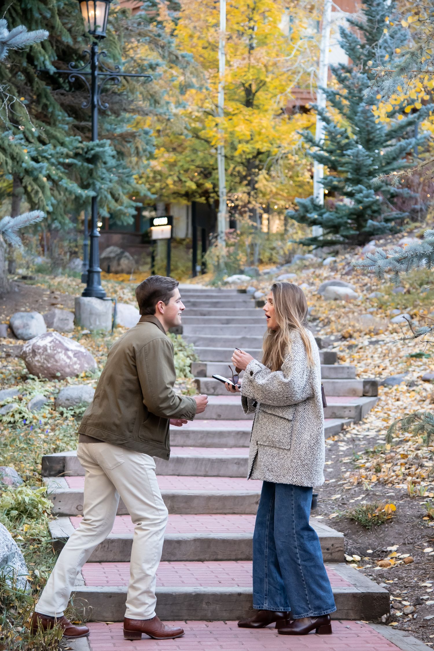 A romantic proposal scene unfolds on outdoor stone steps surrounded by evergreen trees and autumn foliage in Vail Village Colorado.