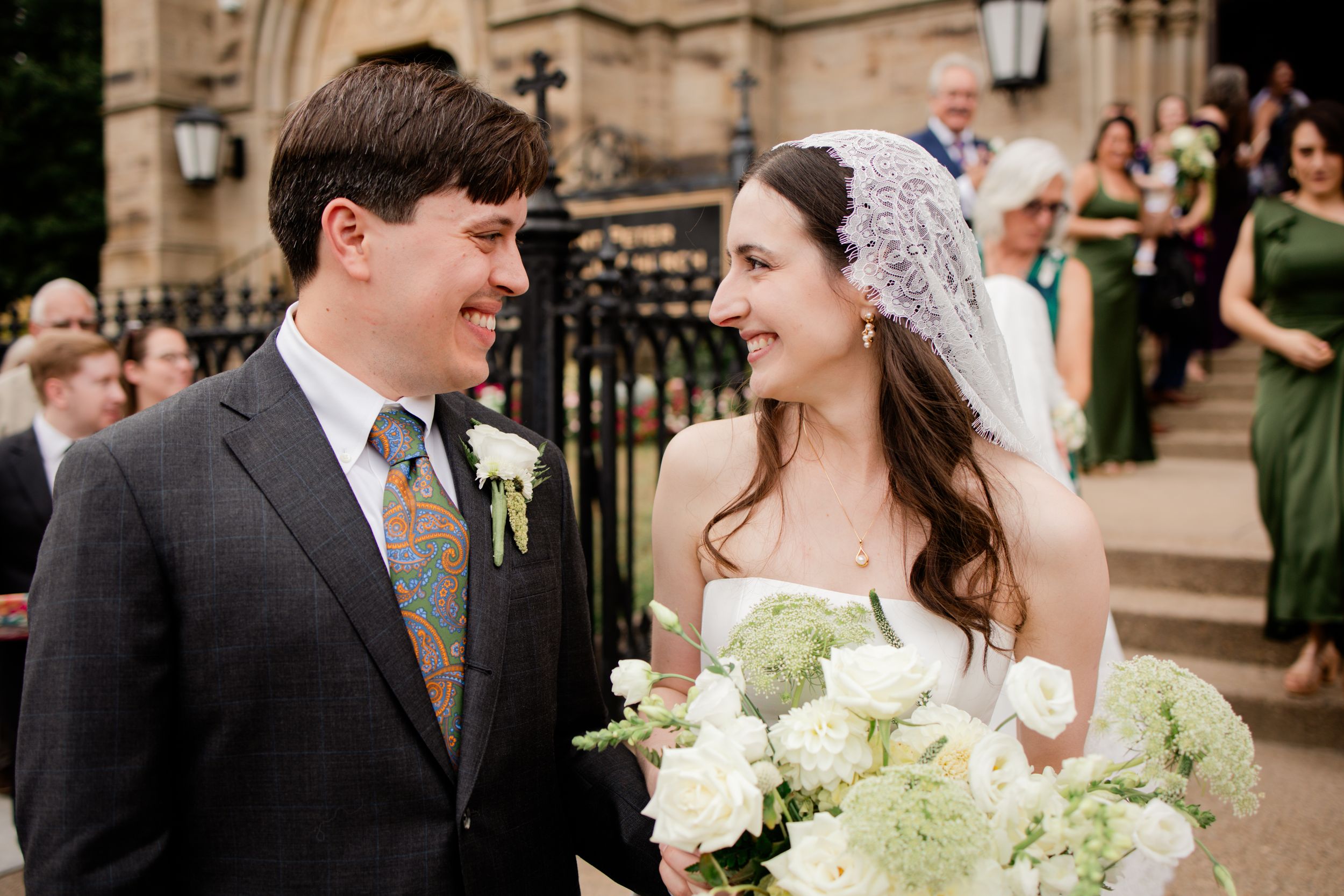 Joyful wedding couple shares a candid moment surrounded by guests outside a historic stone church building.